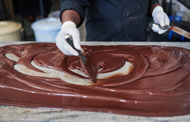 Крупный план работника, использующего лопатки для разбрасывания расплавленного шоколада на стол во время работы на фабрике по производству шоколада
 - Фото, изображение