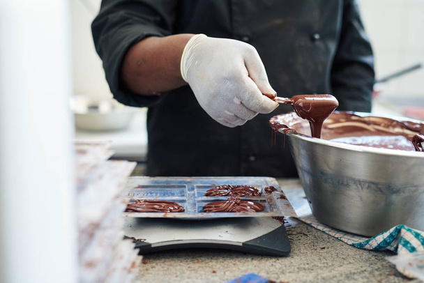 Closeup of a worker standing at a table in an artisanal chocolate making factory pouring melted chocolate into plastic molds - Valokuva, kuva
