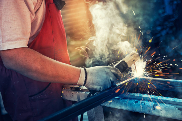Strong man is a welder in T-shirt in a construction gloves, a metal product is welded with a welding machine in the garage, orange sparks fly to the sides - Photo, Image