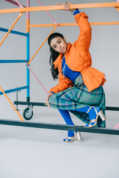fashionable african american female model in orange winter jacket, checkered skirt and blue tights posing near colorful scaffold in studio - Фото, зображення