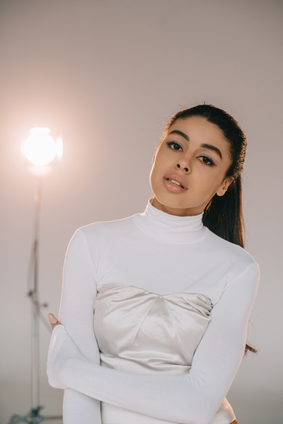 pretty african american girl in white turtle neck looking at camera in studio with spotlight - Zdjęcie, obraz