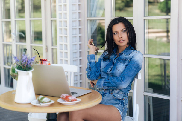 Beautiful attractive woman sits at a table in a cafe, dressed in a denim shirt and shorts, holds her smartphone in her hand, waits for a girlfriend, ordered desserts and cocktails. - Fotografie, Obrázek