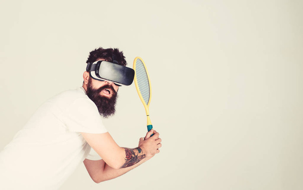 Virtual sport concept. Man with beard in VR glasses beating pitch, grey background. Hipster on busy face use modern technology for sport games. Man gaming tennis in VR headset hitting - Photo, Image