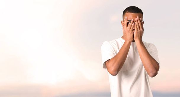 Young african american man covering eyes by hands and looking through the fingers on sunset background - Fotoğraf, Görsel