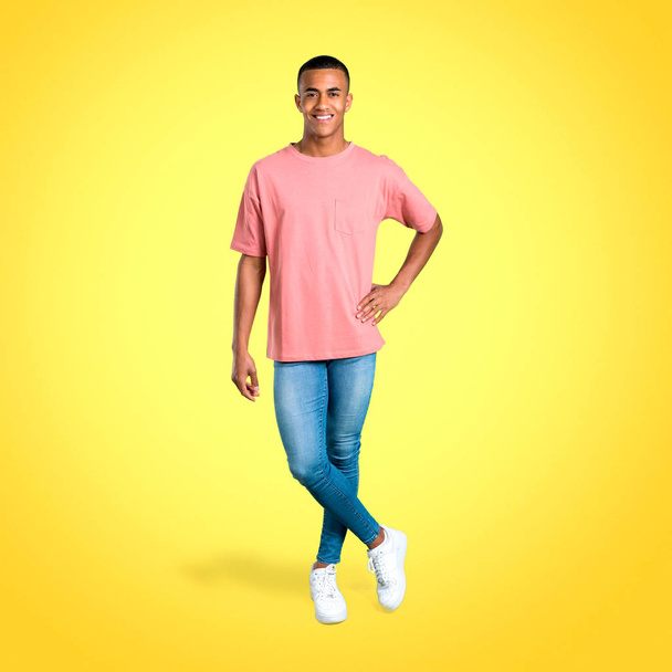 Jeune homme afro-américain debout posant avec les bras à la hanche et riant en regardant vers l'avant sur fond coloré
 - Photo, image