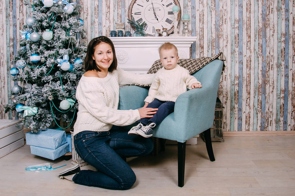 Little boy sitting in armchair near fireplace with mom in the festively decorated room with Christmas tree. Portrait of mother and baby boy - Foto, immagini