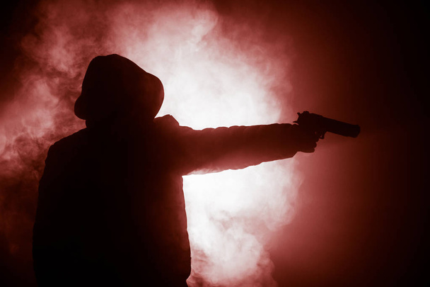 Σιλουέτα του ανθρώπου με πιστόλι έτοιμοι να επιτεθούν στο σκοτάδι τονισμένο ομιχλώδες φόντο ή επικίνδυνο ληστή εκμετάλλευση όπλο στο χέρι. Γυρίσματα τρομοκράτης με το όπλο θέμα ντεκόρ - Φωτογραφία, εικόνα