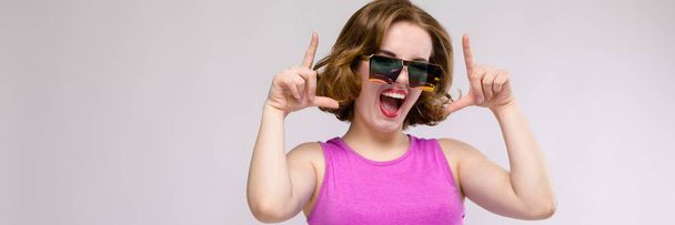 Charming young girl in pink dress on gray background. Cheerful girl in square glasses. The girl points fingers at the top - Foto, Imagem