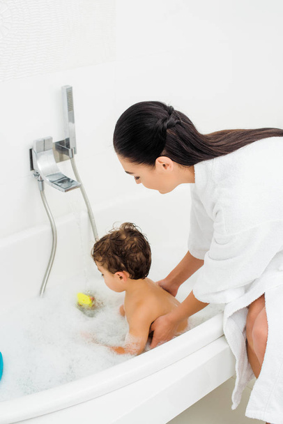 Mom in bathrobe washing son in white bathroom - Foto, afbeelding