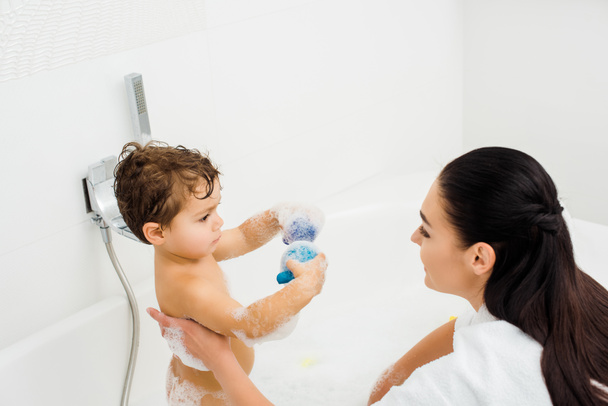 Son showing toys to brunette mother in white bathroom  - Photo, Image