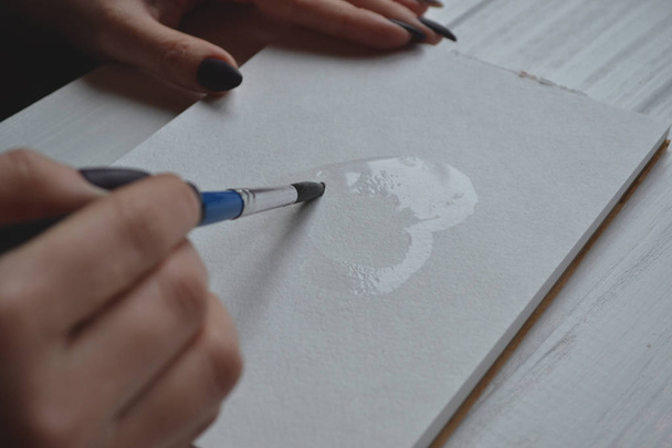 Una donna dipinge in un quaderno. Strumenti per dipingere sulla scrivania in legno bianco. Lavori di pittura
. - Foto, immagini