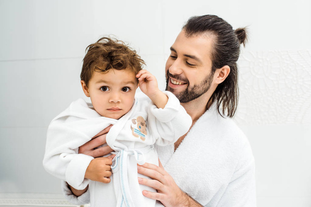 Dad looking at toddler son in white bathrobe and smiling  - Fotoğraf, Görsel