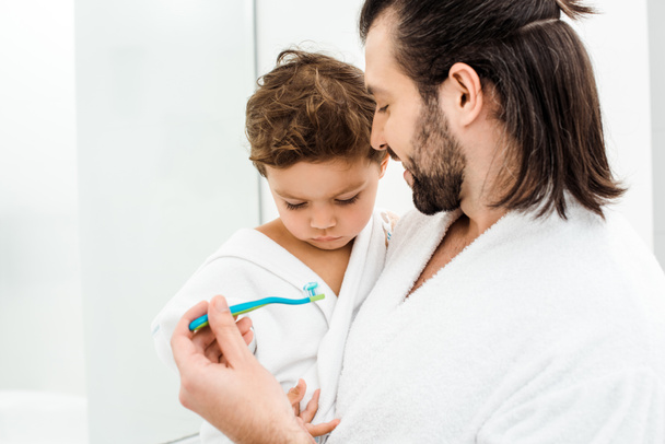 Крупный план отца, показывающего зубную щетку с зубной пастой сыну малыша
  - Фото, изображение
