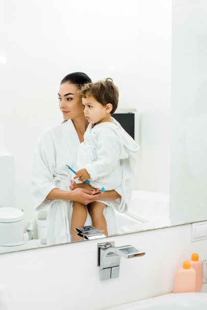 mother and son in white bathrobes brushing teeth together - Fotó, kép