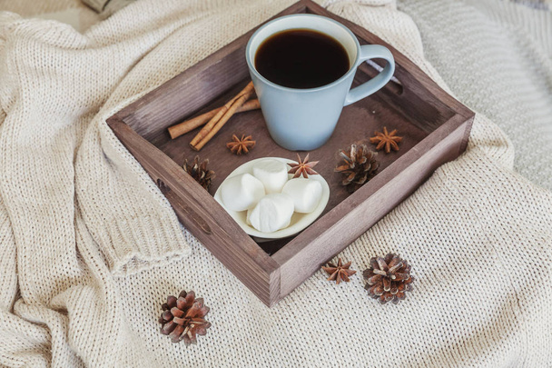 Cup of coffee on rustic wooden tray, sweet marshmallows and warm woolen sweater. Cozy autumn or winter weekend or holidays at home. Fall home decoration with hot drink mug. Hygge morning style concept - Photo, Image