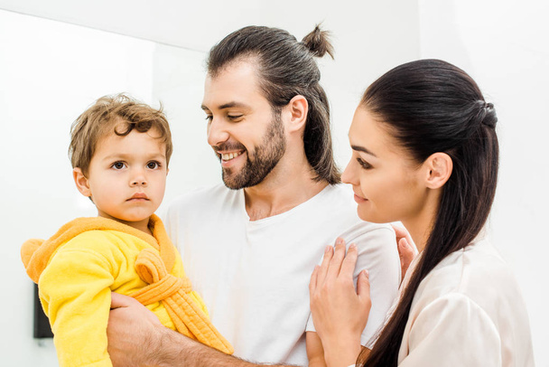 smiling parents holding cute toddler son in yellow bathrobe in bathroom - Photo, Image