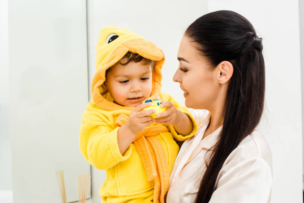 beautiful woman holding son in yellow bathrobe with rubber toy - Photo, Image