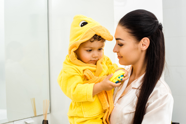 jovem mãe segurando bonito filho em roupão amarelo com brinquedo
 - Foto, Imagem