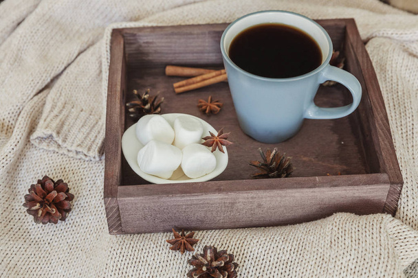 Cup of coffee on rustic wooden tray, sweet marshmallows and warm woolen sweater. Cozy autumn or winter weekend or holidays at home. Fall home decoration with hot drink mug. Hygge morning style concept - Photo, Image