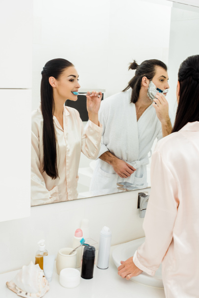 beautiful woman brushing teeth and handsome man shaving in front of mirror in bathroom  - Фото, зображення