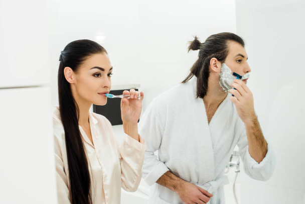 Beautiful woman cleaning teeth while husband shaving beard - Photo, Image