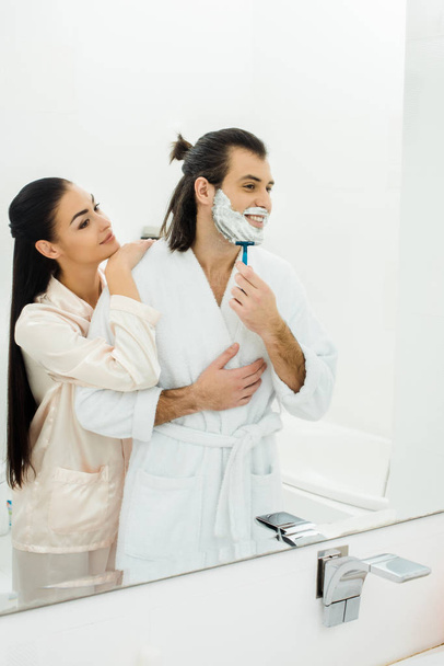Pretty woman embracing  husband while man shaving in front of mirror - Foto, imagen