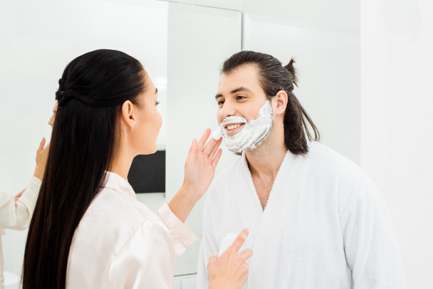 Hombre guapo sonriendo mientras que la mujer que aplica espuma de afeitar en la cara del hombre
 - Foto, imagen