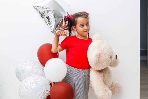 Портрет улыбающейся веселой девушки с плюшевым мишкой и красочными воздушными шарами
 - Фото, изображение