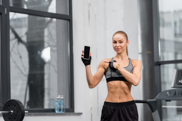 attractive athletic sportswoman pointing on smartphone with blank screen in gym - Photo, Image