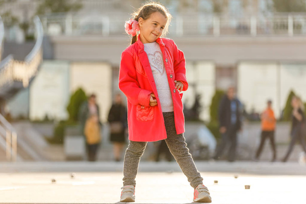Little girl in red coat walking in sunny city street - Фото, изображение
