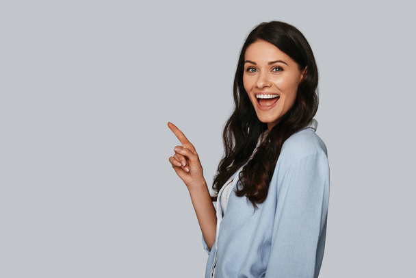 Happy young woman looking at camera and pointing finger on left side, copy space on grey background - Photo, image