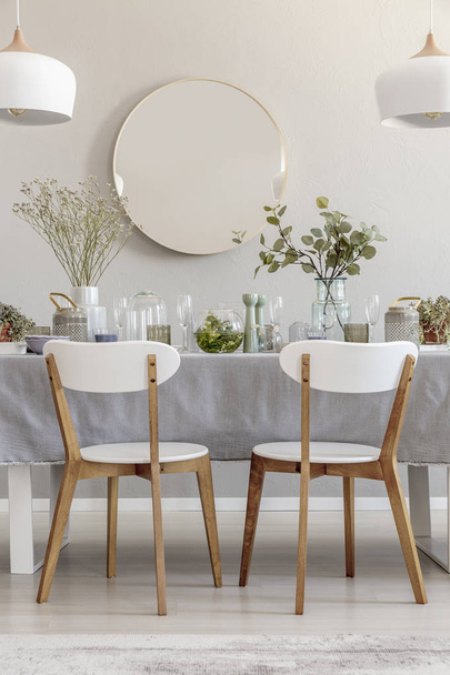 Two white chairs standing by the table with fresh plants and glass vessels in the photo of bright dining room interior with mirror on the wall - Foto, Imagem