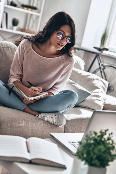 Beautiful young woman in glasses studying while sitting on sofa at home and writing in texbook with pen  - Photo, image