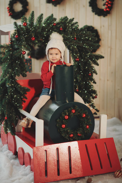 Happy little child girl in winter knitted hat sitting on a Christmas wooden train, decorated with Christmas tree branches and balls - Fotó, kép