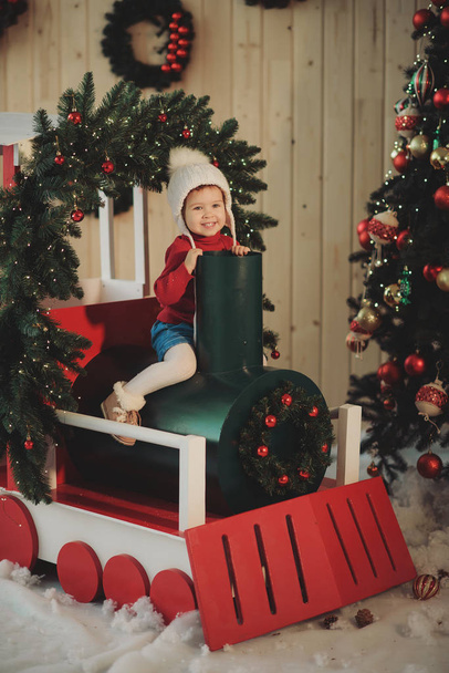 Happy little child girl in winter knitted hat sitting on a Christmas wooden train, decorated with Christmas tree branches and balls - Foto, Bild