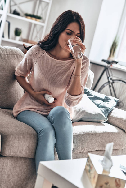 female Periods, Young sad woman suffering from stomachache pain while sitting on sofa at home and drinking water from glass          - Фото, изображение