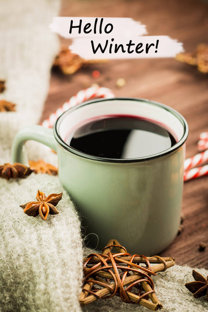 Winter theme. Hot steaming cup of glint wine with spices, cinnamon, anise, red candies, pepper and gray scarf on wooden background with text Hello Winter. View from above. - Photo, Image