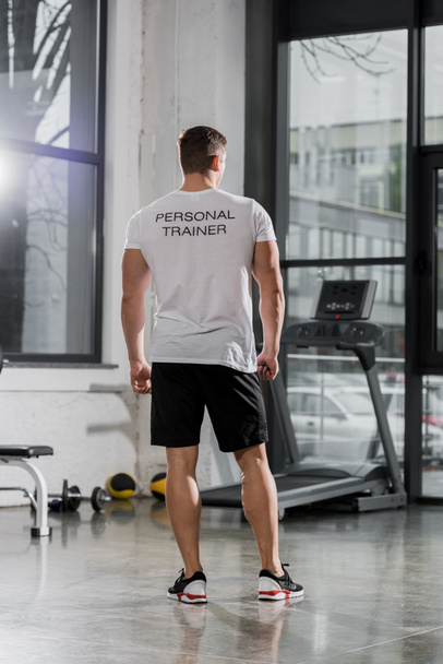 back view of athletic bodybuilder in shirt with text personal trainer standing in gym - Foto, Imagen