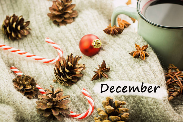 Christmas hot steaming cup of glint wine with spices, cinnamon, anise, cookies in a shape of star, red candies, fir cones, pepper and gray scarf on wooden background with text December. - Φωτογραφία, εικόνα