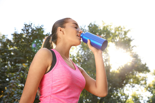 Young sporty woman drinking from water bottle in park on sunny day - Фото, изображение