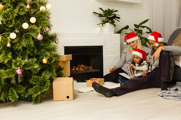 Rodina v červené santa klobouky s přítomné sedící na podlaze vánoční stromeček v obýváku  - Fotografie, Obrázek