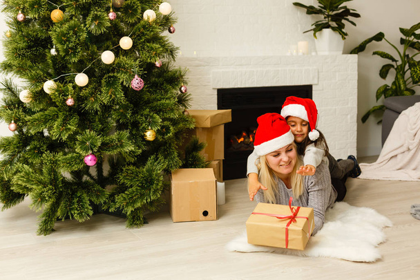 мать с дочерью в красных шляпах Санты с подарком, лежащим на полу возле елки в гостиной
  - Фото, изображение