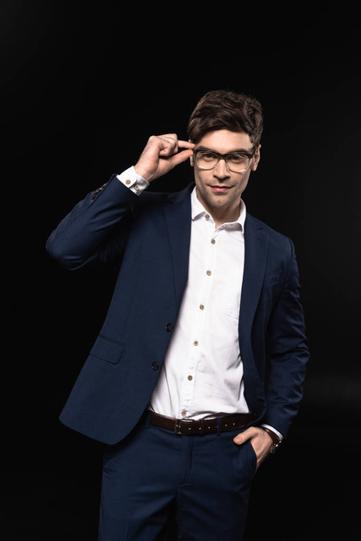 handsome young businessman in stylish suit and eyeglasses looking at camera isolated on black - Φωτογραφία, εικόνα