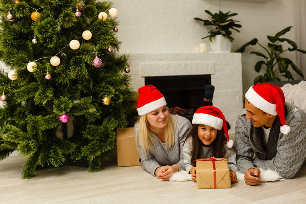 Family in red santa hats with present lying on floor near Christmas tree at living room  - Фото, зображення