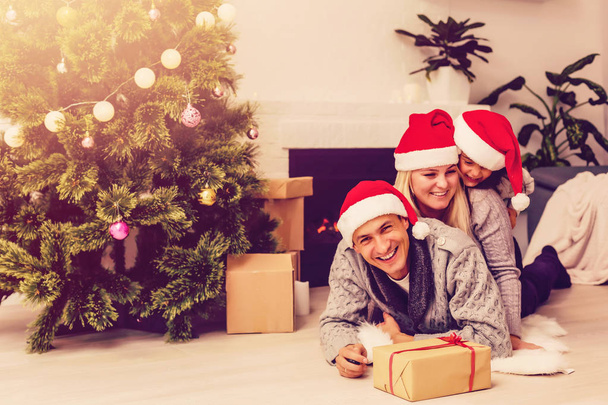 Family in red santa hats with present lying on floor near Christmas tree at living room  - 写真・画像