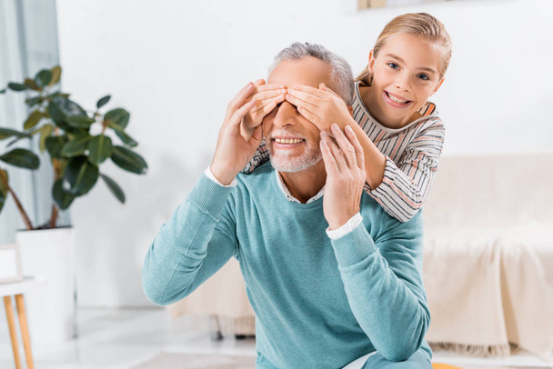 adorable little kid looking at camera and covering eyes of smiling grandfather at home - Photo, image