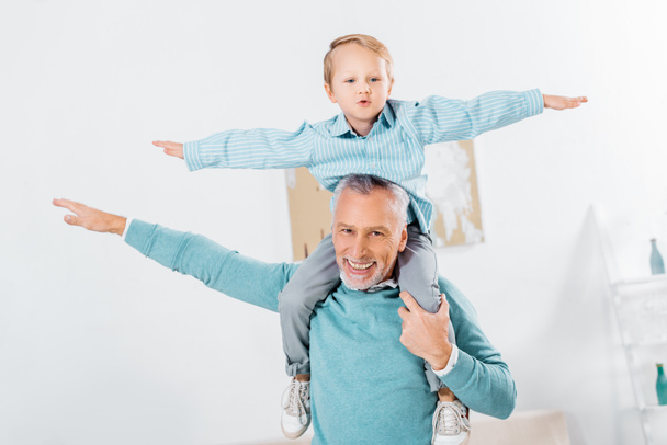 child sitting on happy grandfather shoulders and imitating plane at home - Photo, Image