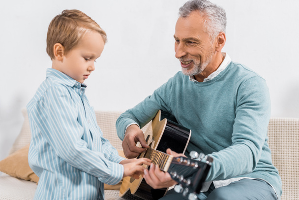 smiling middle aged man teaching grandson playing on acoustic guitar at home - Foto, immagini