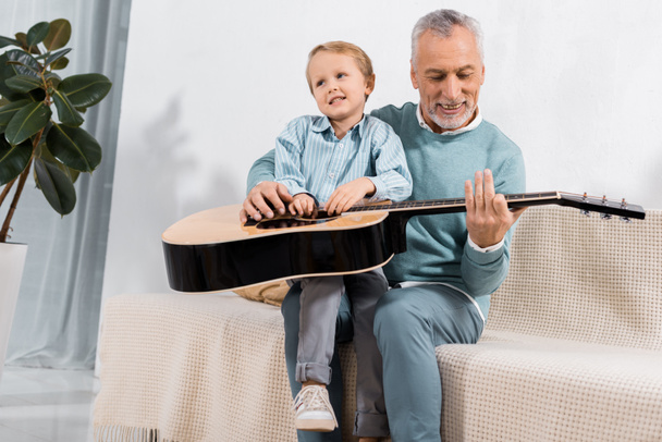 cheerful grandfather playing with grandson on knees playing on acoustic guitar at home - Photo, image