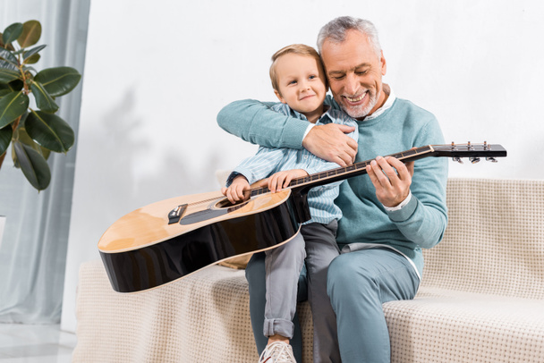selective focus of laughing grandfather playing with grandson on knees playing on acoustic guitar at home - Photo, image